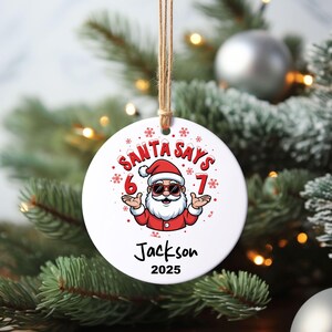 May include: A white Christmas ornament with a Santa Claus graphic. The ornament reads "SANTA SAYS 6 7" and includes the name "Jackson" and the year "2025". The ornament is hanging from a natural twine string.