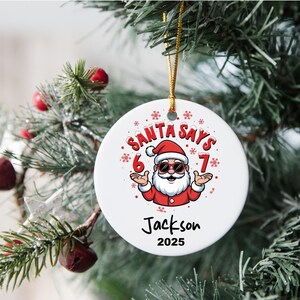 May include: A white ceramic Christmas ornament with a cartoon Santa Claus design. The ornament features the text "SANTA SAYS" above the Santa illustration, with "6" and "7" on either side. The name "Jackson" and the year "2025" are below.