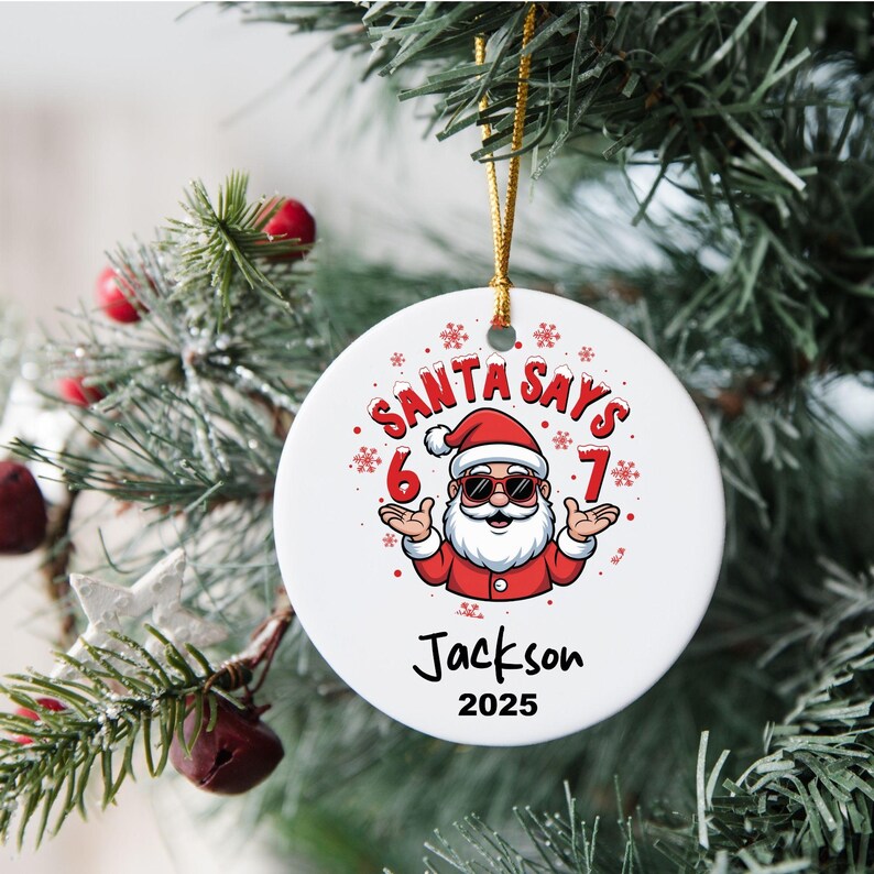 May include: A white ceramic Christmas ornament with a cartoon Santa Claus design. The ornament features the text "SANTA SAYS" above the Santa illustration, with "6" and "7" on either side. The name "Jackson" and the year "2025" are below.