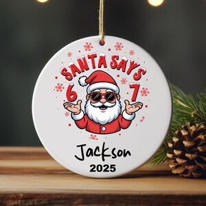 May include: White Christmas ornament with a cartoon Santa Claus wearing sunglasses, a red suit, and a white beard. The text "SANTA SAYS 6 7" is above the Santa illustration. Below is the name "Jackson" and the year "2025".