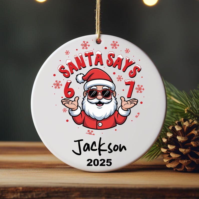 May include: White Christmas ornament with a cartoon Santa Claus wearing sunglasses, a red suit, and a white beard. The text "SANTA SAYS 6 7" is above the Santa illustration. Below is the name "Jackson" and the year "2025".