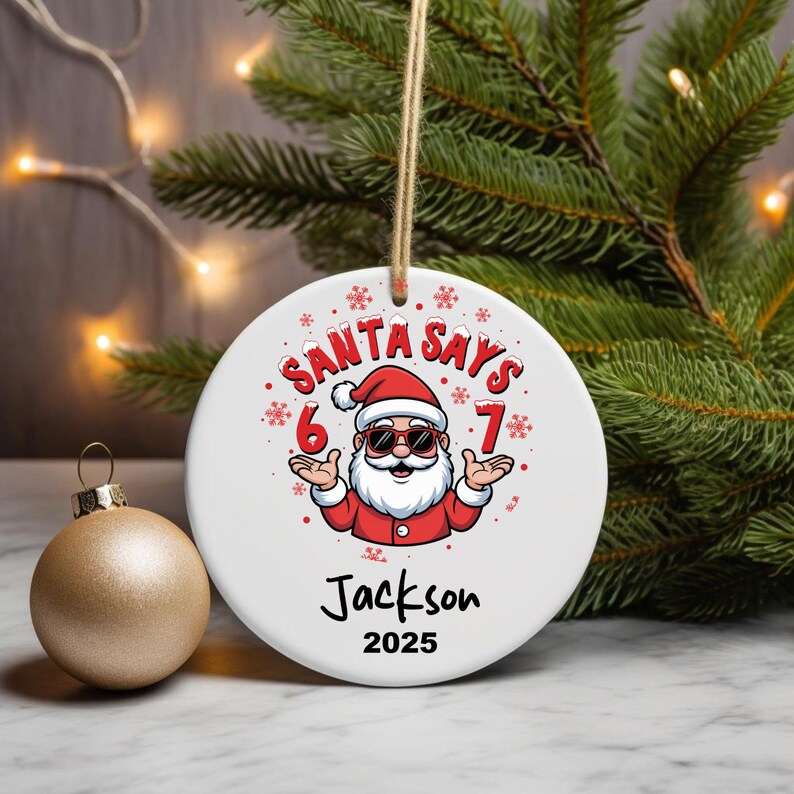 May include: A white ceramic Christmas ornament with the text "SANTA SAYS" and a cartoon Santa Claus wearing sunglasses. The ornament also includes the name "Jackson" and the year "2025". A gold-colored ornament is in the background.