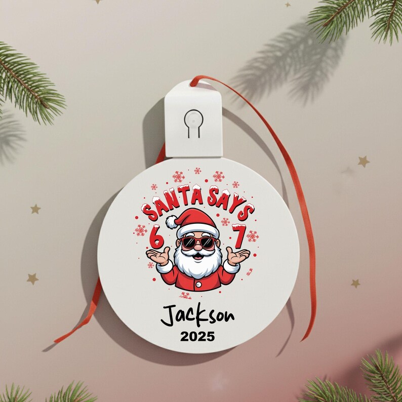 May include: A white Christmas ornament with a red ribbon features a cartoon Santa Claus wearing sunglasses. The text "SANTA SAYS 6 7" is above the Santa illustration, with the name "Jackson" and the year "2025" below.