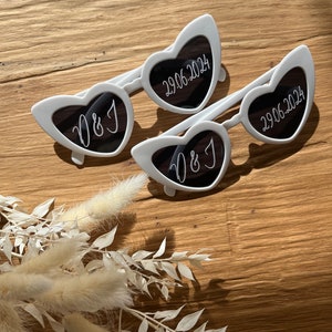 May include: Two pairs of white heart-shaped sunglasses. One pair has the initials "D&J" on the lenses, and the other pair has the date "29.06.2024" on the lenses.