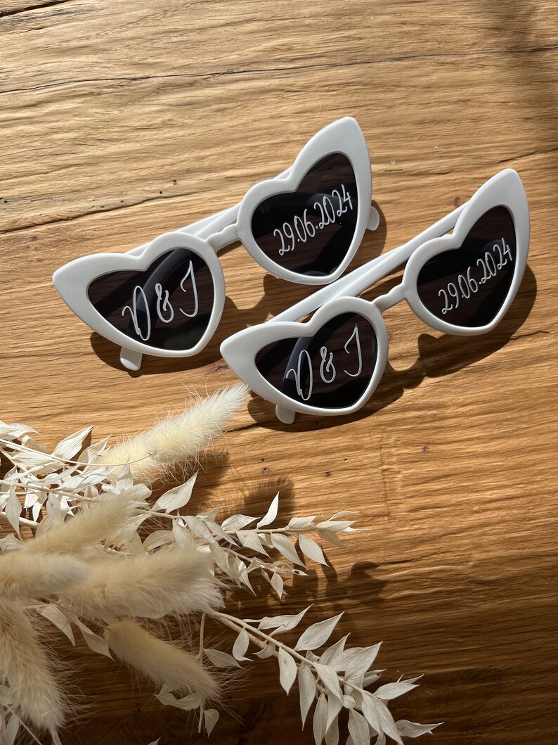 May include: Two pairs of white heart-shaped sunglasses. One pair has the initials "D&J" on the lenses, and the other pair has the date "29.06.2024" on the lenses.