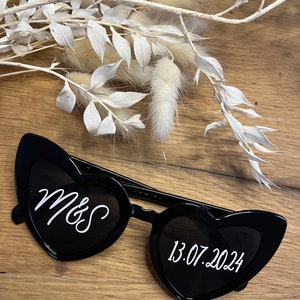 May include: Black heart-shaped sunglasses with white text. The left lens says "Mrs" and the right lens says "13.07.2024".