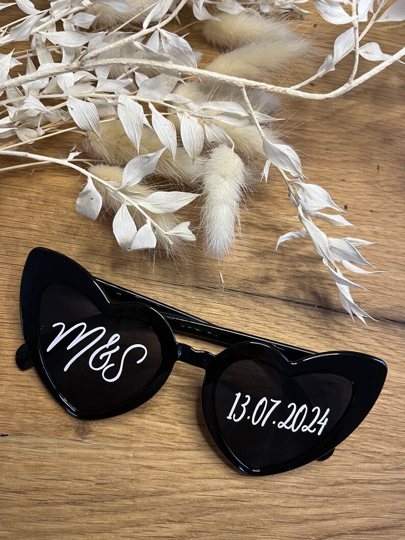 May include: Black heart-shaped sunglasses with white text. The left lens says "Mrs" and the right lens says "13.07.2024".