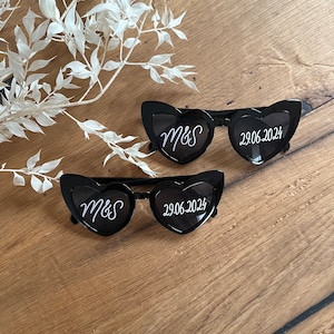 May include: Two black heart-shaped sunglasses with white text. The left sunglass says "M&S" and the right sunglass says "29.06.2024".