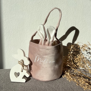 May include: A blush pink velvet Easter basket with white bunny ears peeking out. The name "Melina" is written on the side. A white wooden bunny with a heart cutout and twine bow sits next to the basket, suggesting an Easter theme.