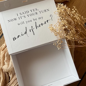 May include: White gift box with the text "I SAID YES, NOW IT'S YOUR TURN will you be my maid of honor?" in black script. The box is open, revealing an empty interior. Dried flowers are placed near the box.