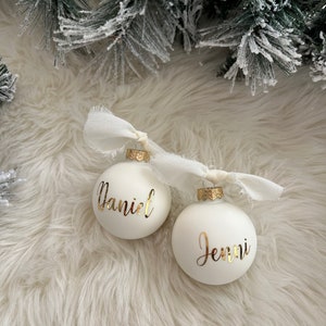 May include: Two white Christmas ornaments with gold lettering. One ornament says "Daniel" and the other says "Jenni".