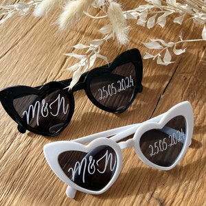 May include: Two pairs of heart-shaped sunglasses, one black and one white. Both pairs have the text "M&H" and "25.05.2024" written on the lenses.