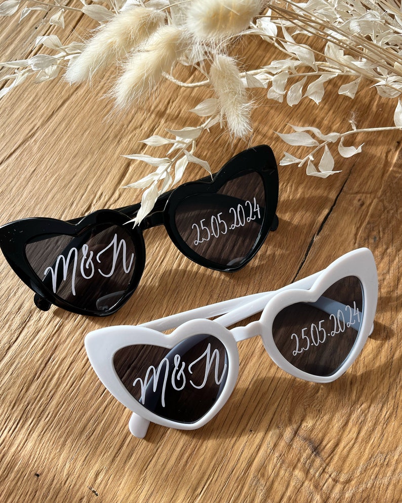 May include: Two pairs of heart-shaped sunglasses, one black and one white. Both pairs have the text "M&H" and "25.05.2024" written on the lenses.