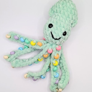 May include: A mint green crocheted plush squid toy featuring a soft, textured yarn body, two black plastic safety eyes, and a stitched black mouth. The eight tentacles are adorned with various colorful plastic beads in pastel shades.