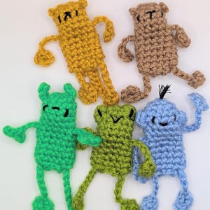 May include: Five small, flat, crocheted figures in yellow, tan, bright green, olive green, and light blue. Each figure has a rectangular body, thin limbs with rounded ends, and simple embroidered facial features in black yarn.
