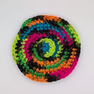 May include: A circular crochet coaster featuring a vibrant spiral pattern. The yarn colors include bright orange, lime green, hot pink, electric blue, and black, worked in a continuous round stitch that radiates from the center to the outer edge.