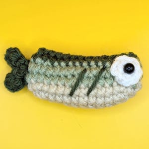 May include: A small, handmade crochet fish toy featuring a gradient of dark green, light green, and cream yarn. The fish has a dark green tail, two dark green stitched gill lines, and a white crocheted eye with a black plastic bead center.