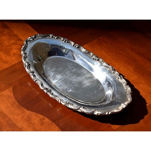 May include: Oval silver serving tray with ornate, scalloped edges. The tray has a reflective surface and is likely made of silver or a silver-plated material. The design is classic and elegant, suitable for serving food or displaying items.