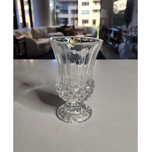 May include: A clear glass vase with a diamond pattern and a wide base. The vase is empty and has a classic design.