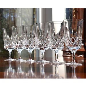 May include: A set of six clear crystal champagne flutes. Each glass features a diamond-cut pattern on the bowl and a slender stem. The glasses are arranged in a row, reflecting light and creating a sparkling effect.