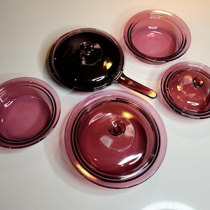 May include: Set of five purple glass cookware pieces, including a skillet with a handle and four lids. The lids have a raised, textured design.