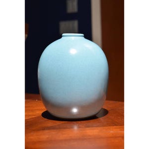 May include: A light blue ceramic vase with a rounded shape. The vase is smooth and has a glossy finish.