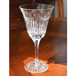 May include: A clear crystal wine glass with a long stem and a wide, patterned base. The bowl of the glass features a starburst design and vertical cuts. The glass is sitting on a wooden surface.