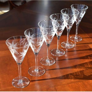 May include: Six clear glass wine glasses with etched leaf designs stand in a row on a wooden table. The glasses are arranged from largest to smallest, with the largest glass at the top of the image.