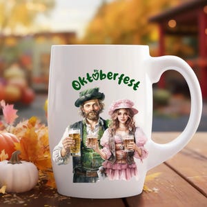 May include: White ceramic mug with a handle, featuring a festive Oktoberfest design. The mug depicts a man and woman in traditional German attire, holding beer steins. The text "Oktoberfest" is written in green lettering.