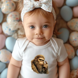 May include: A baby wearing a white onesie with a graphic print of a hairy creature with a finger to its lips. The baby is surrounded by pastel blue and pink Easter eggs.