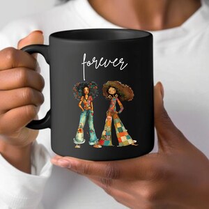 May include: Black ceramic mug featuring a colorful illustration of two women in retro outfits. The mug has the word "forever" in white script. The women are wearing bell-bottom jeans and patchwork pants.