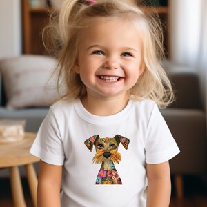 May include: A young child wearing a white t-shirt with a colorful, patterned dog illustration. The dog has a brown and white face with a floral design.