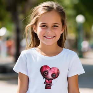 May include: A white t-shirt with a cartoon heart design. The heart is red with dark details and large eyes. The design is centered on the shirt. The person wearing the shirt is smiling.