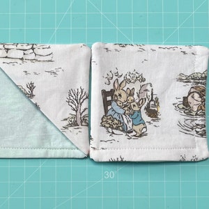 Puede incluir: Dos posavasos de tela con fondo blanco y borde azul claro. Un posavasos presenta un diseño de Peter Rabbit con una familia de conejos. El otro posavasos tiene una tela azul claro en un lado y un diseño de Peter Rabbit en el otro.