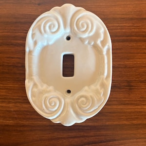 Peut inclure: Un cache-interrupteur en céramique blanche avec un motif tourbillonnant orné. Le cache a une forme ovale avec une ouverture rectangulaire pour l'interrupteur et deux petits trous de montage. Fond en bois foncé.