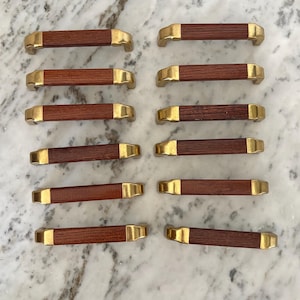 May include: Twelve cabinet pulls with a dark brown wooden centre and gold-coloured metal ends. The pulls are arranged on a marble-effect surface. Each pull is rectangular in shape.