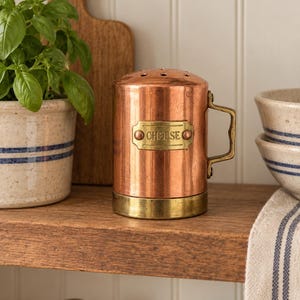 May include: A cylindrical copper cheese shaker with a brass base, a brass handle, and a brass label plate featuring the word CHEESE in embossed lettering. The top of the shaker is domed and perforated with small holes for dispensing grated cheese.