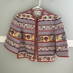 May include: A cropped jacket featuring a multi-coloured floral and geometric pattern in shades of red, blue, yellow, and white. The garment has three-quarter length sleeves, a round neckline, and a front opening. A label reads 'Vicki Lynn London'.