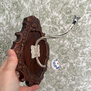 May include: A decorative wall hook with a dark brown, ornate wooden backplate. The hook has a white, curved metal arm and a white ceramic knob with blue floral designs and a red dot.