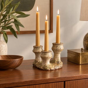 May include: A ceramic triple candle holder with a speckled, off-white glaze and a textured, organic base. Three tall, slender, light yellow taper candles are inserted into the holders, each with a lit flame, creating a warm, vertical glow.