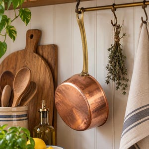 May include: A round copper saucepan with a long, curved brass handle hanging from a metal hook. The pan features a polished metallic finish and is suspended against a white vertical paneled wall, positioned next to a bundle of dried herbs.