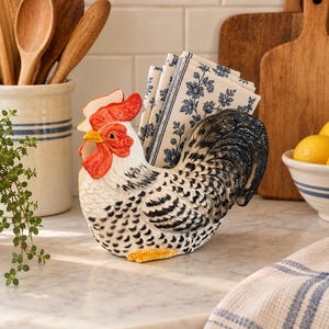 Peut inclure: Un porte-serviettes en céramique en forme de coq avec un corps blanc aux détails noirs tachetés, une crête et des barbillons rouge vif, un bec jaune et une queue gris foncé. Il contient des serviettes blanches à motif floral bleu.