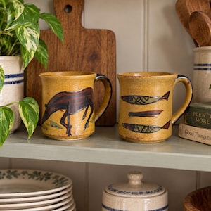 May include: Two ceramic mugs with a speckled mustard yellow glaze. The left mug features a dark brown hand-painted horse, while the right mug displays two stylised fish with cross-hatched patterns and a horizontal line between them.