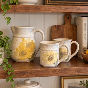 May include: A ceramic pitcher and two matching mugs featuring a speckled off-white finish and hand-painted yellow sunflower designs. Each piece has a sturdy handle and a textured surface, showcasing a consistent floral motif across the set.