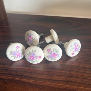 Peut inclure: Un ensemble de cinq boutons de placard ronds en céramique blanc cassé, chacun orné d'un motif floral avec des fleurs roses et violettes et des feuilles vertes. Les boutons ont une finition lisse et brillante et incluent des vis en métal.