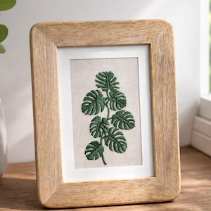 Peut inclure: Une broderie encadrée représentant une plante de monstera verte. L'œuvre est placée sur un fond neutre dans un passe-partout blanc, encadrée dans un cadre en bois clair. Le cadre a des coins arrondis.