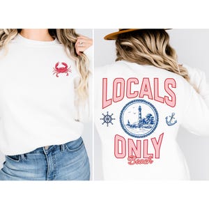 May include: A white crewneck sweatshirt featuring a small red crab graphic on the front left chest. The back displays a large circular nautical design with a lighthouse, a ship's wheel, an anchor, and the text LOCALS ONLY Beach in pink.