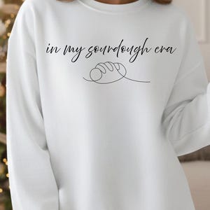May include: White sweatshirt with the phrase "in my sourdough era" in black script. A line drawing of a loaf of bread is below the text. The sweatshirt has a crew neck and long sleeves.