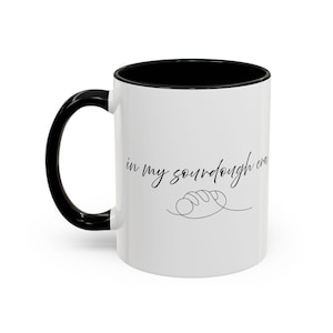 May include: White ceramic mug with a black handle and interior. The mug features the text "in my sourdough era" in a cursive font, along with a line drawing of a loaf of bread. A perfect gift for a baker.