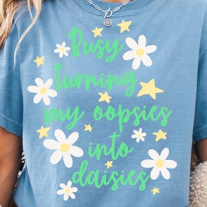 May include: A blue t-shirt featuring the green text Busy turning my oopsies into daisies, surrounded by white daisy flowers with yellow centers and small yellow stars. The shirt has a crew neckline and short sleeves, showcasing a graphic print.